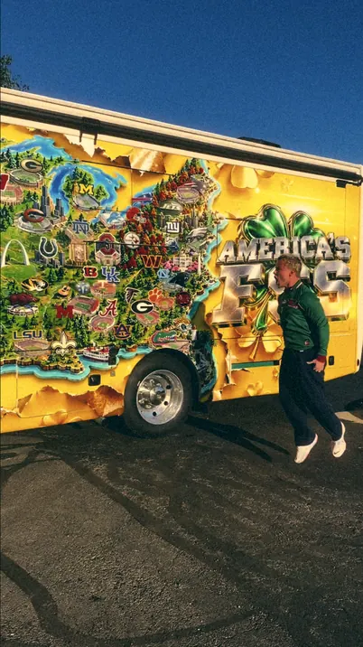Tiki mid-jump beside his yellow tour bus, wrapped in an illustrated map of American stadiums with AMERICA'S FSS on the side